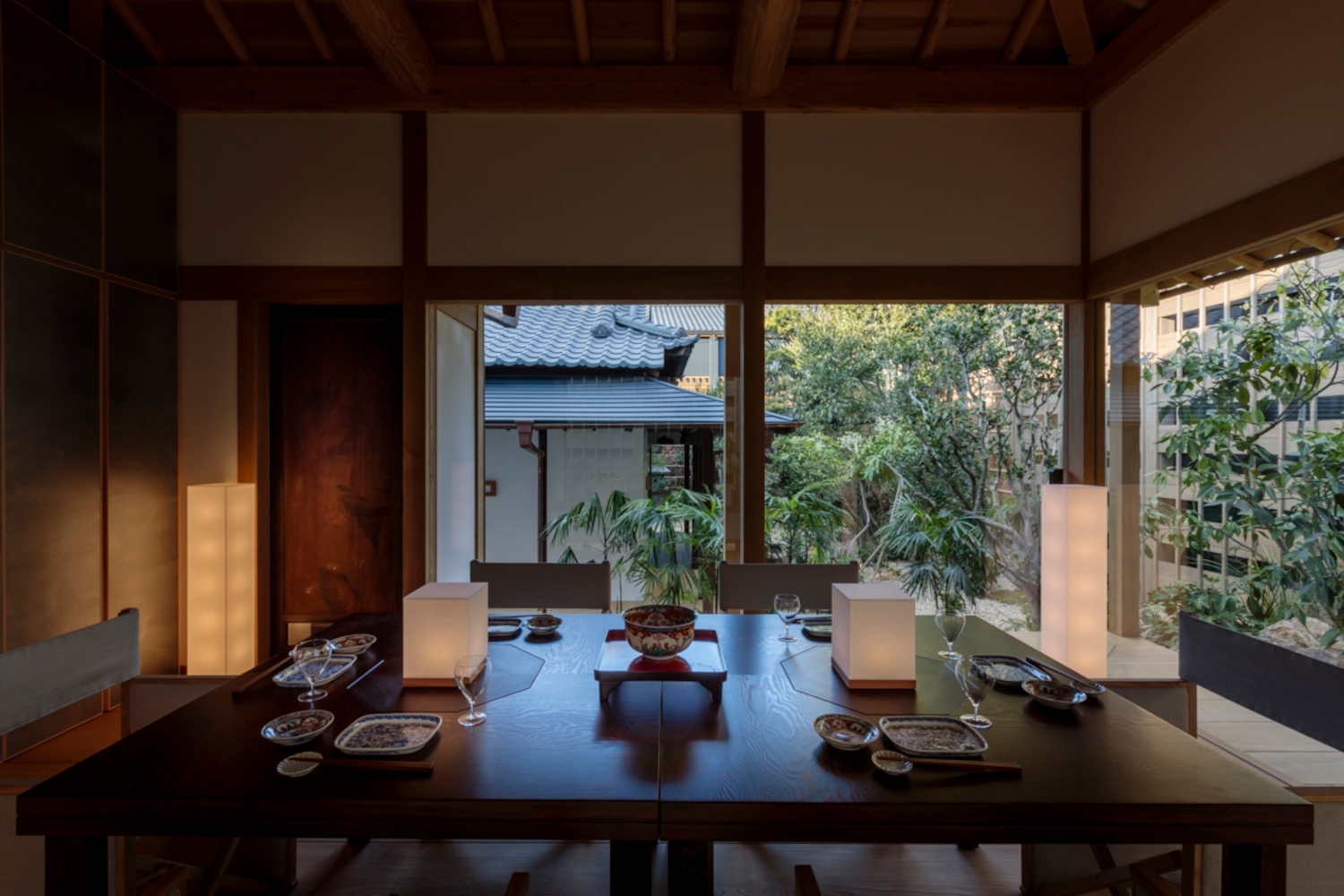 Interior of a dining table with paper lamps set for a meal, and in the background large windows overlooking a central courtyard with Zen garden.