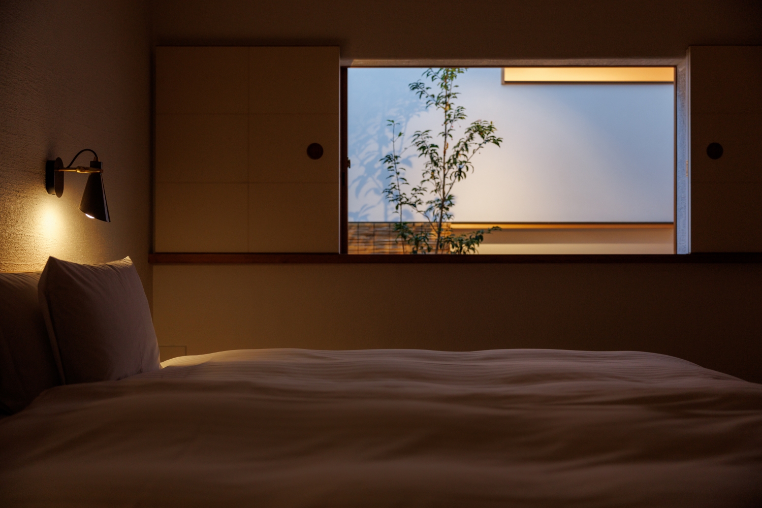 Interior bedroom with a white, minimalist bed and a plain screen window looking out to a zen courtyard with a single small tree.