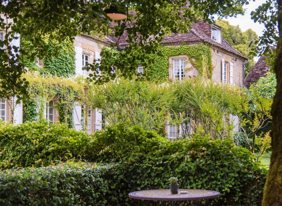 Exterior view of a stone manor house covered in ivy with green hedges and a small table in the foreground.