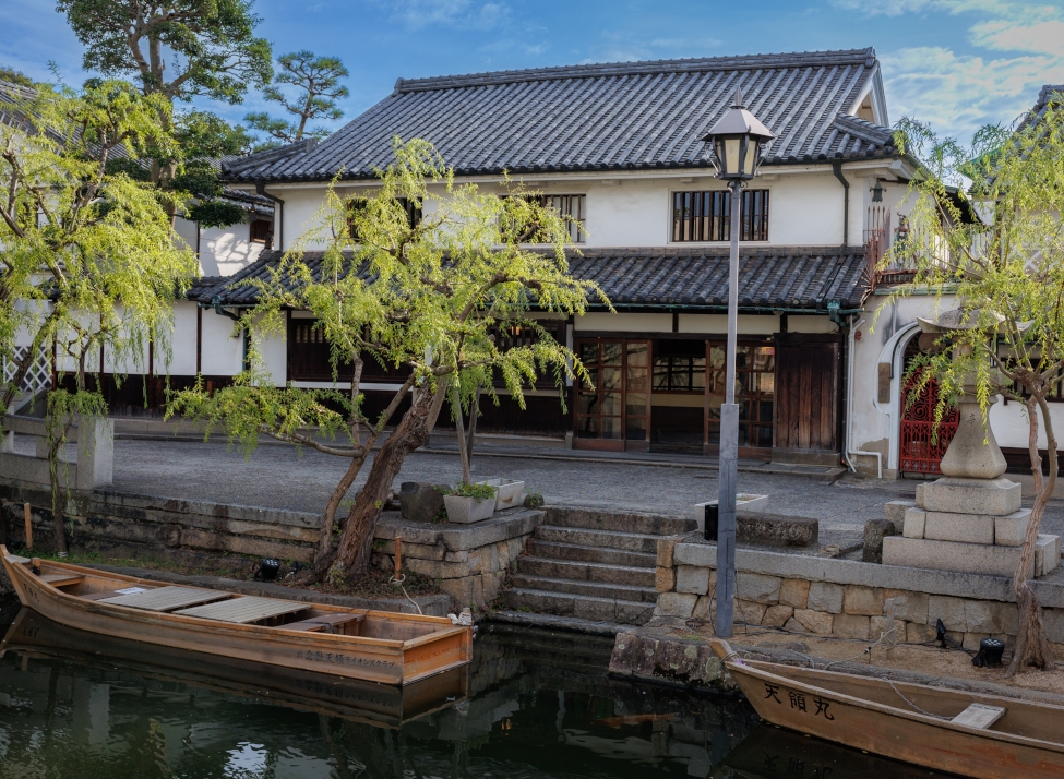 Exterior of a traditional Japanese tile-roofed building along a canal with wooden canoes and willow trees and an old street lamp.