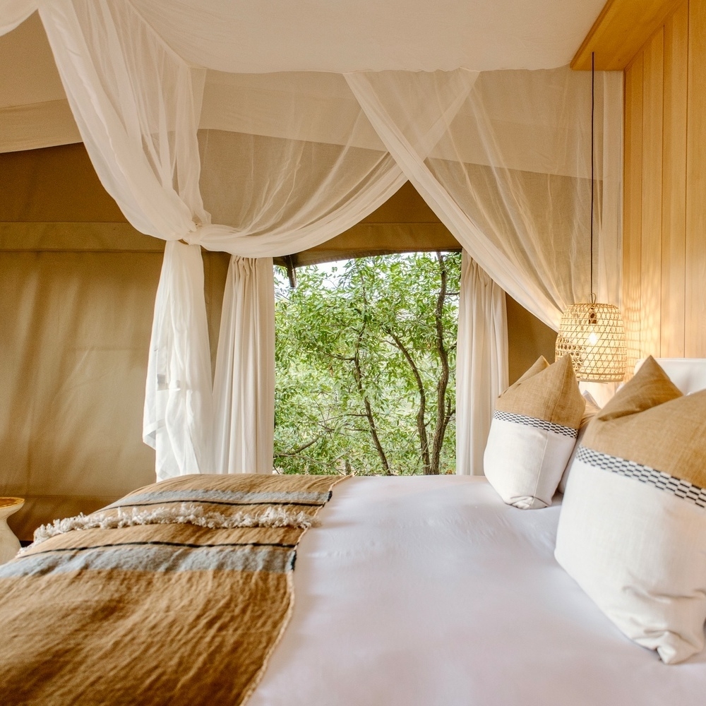 Inside a luxury safari tent showing a bed with a white mosquito net and a view of green trees outside.