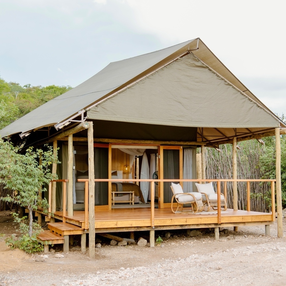 A large canvas safari tent on a wooden platform with two lounge chairs on the front deck.