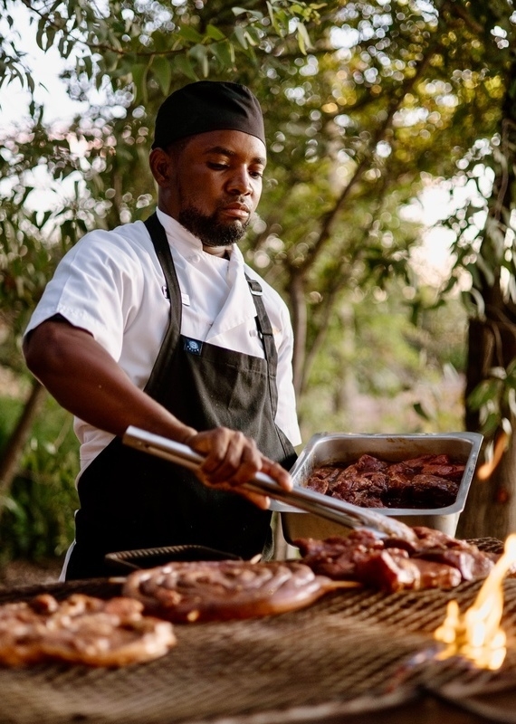 A chef grilling various cuts of meat over an open flame on an outdoor barbecue.