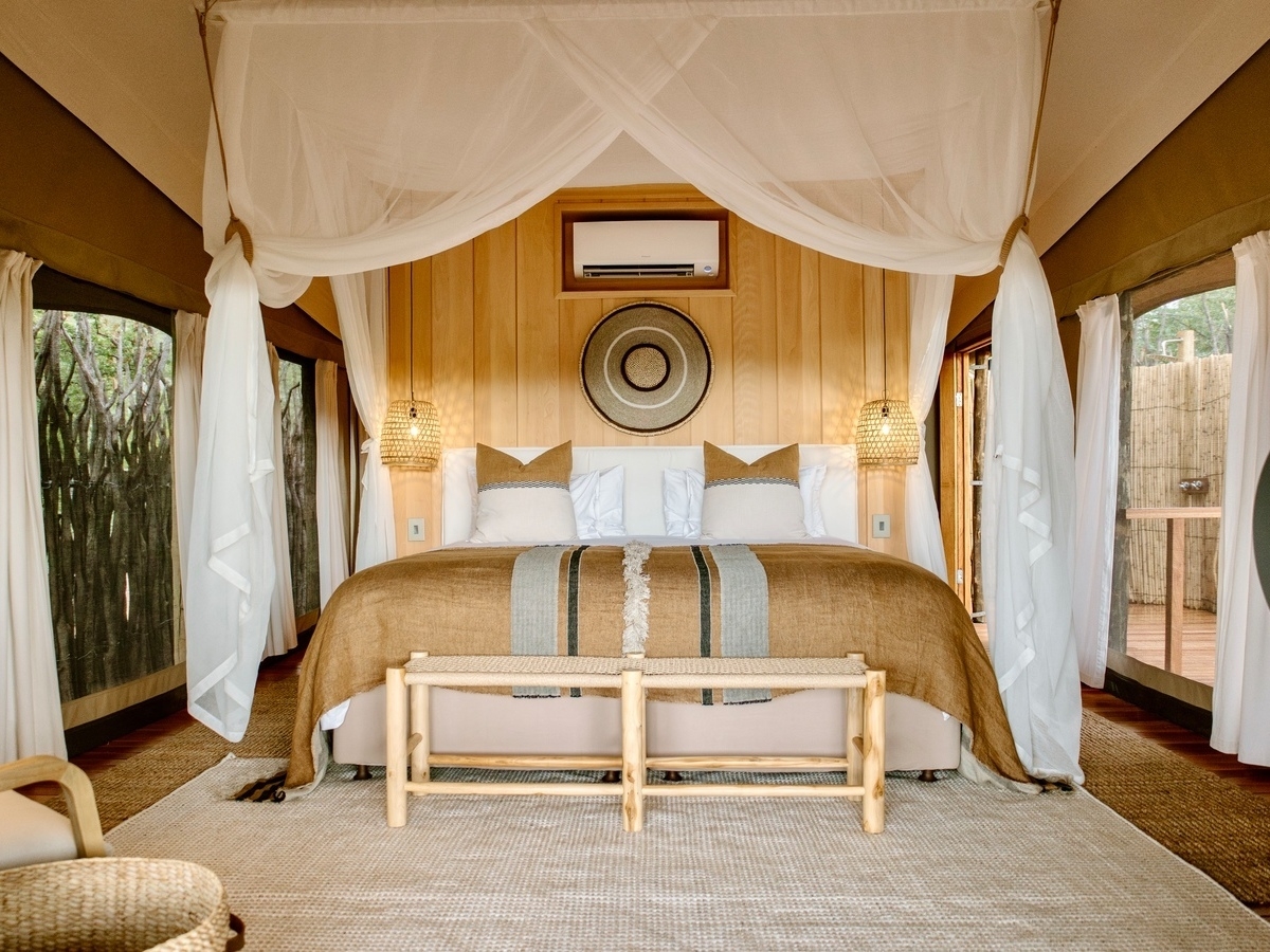 Interior of a safari tent showing a bed centered under a draped mosquito net with symmetric lighting and decor.