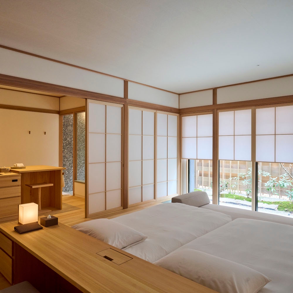 Interior of a bedroom with a minimalist bed with white bedding, light wood frame and white walls with light wood edging and accents, and large windows with paper screen panels.