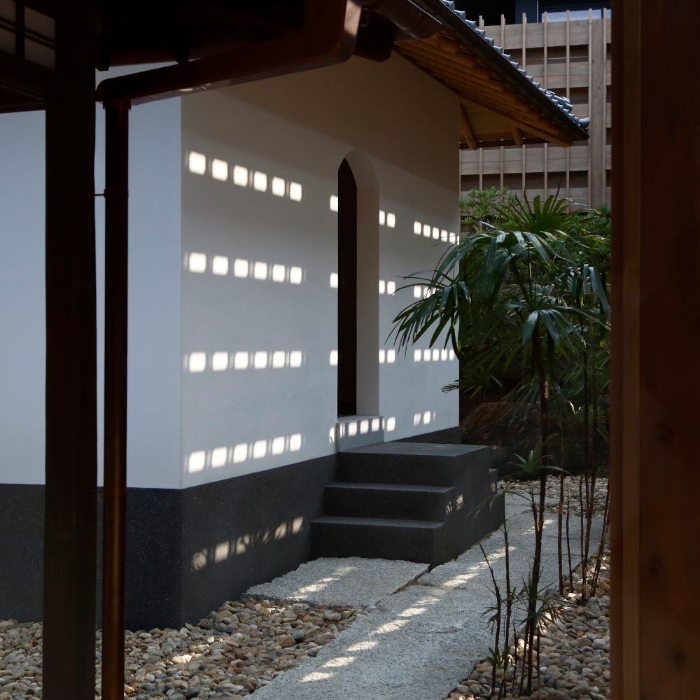 Exterior of an entryway with a rounded door and paved path surrounded by stones, entering into a white-washed building with tile roof.
