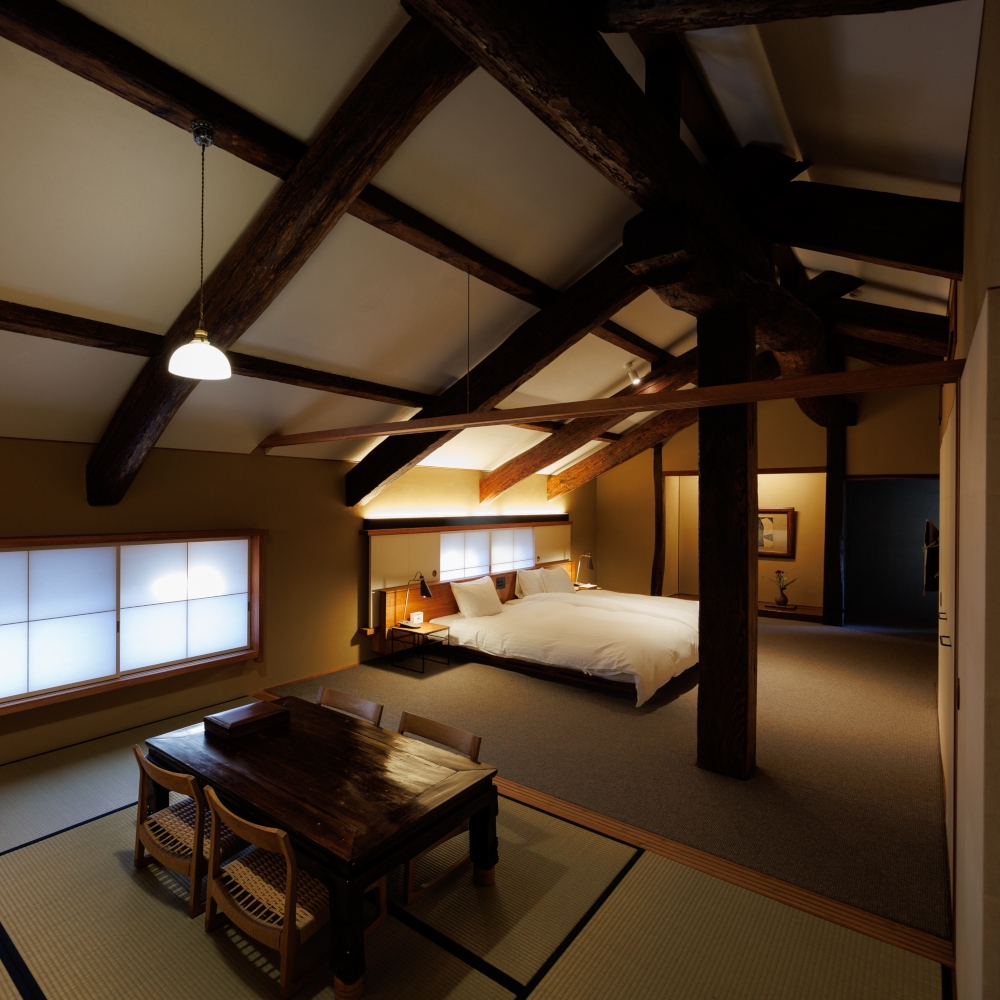 Interior of a ryokan bedroom with a floor-seated tatami table in the foreground and a western-style bed, and high ceilings with wooden beams.