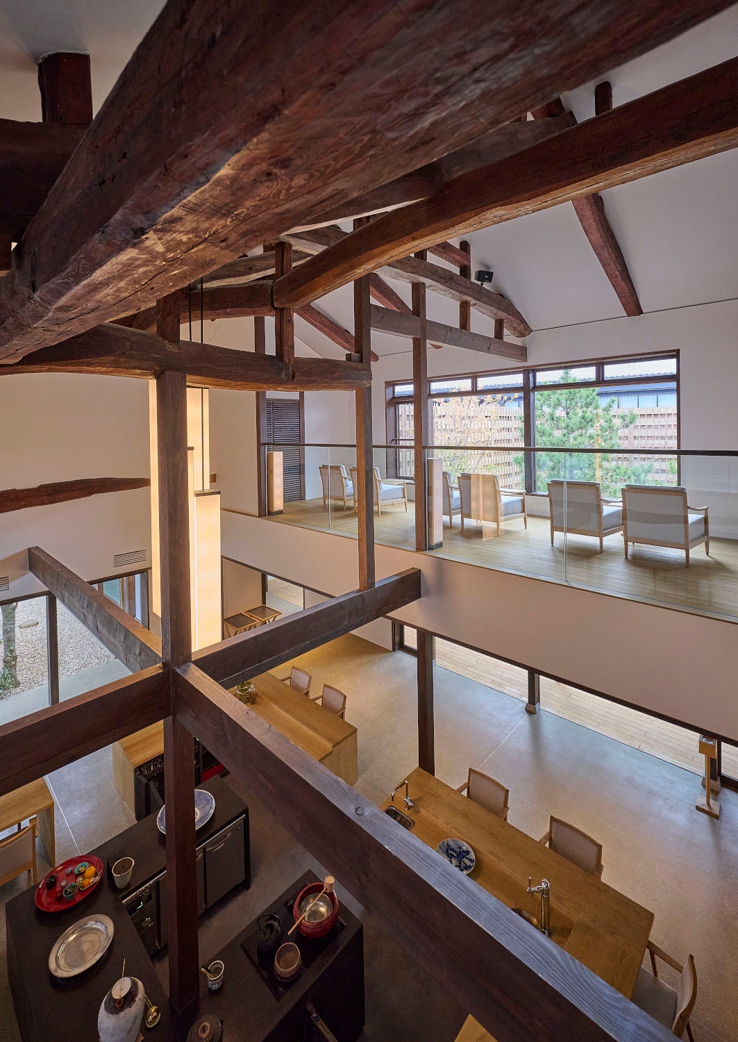 Interior shot of a mezzanine level lounge with several chairs facing a long picture window, and below a lower ground floor with table and dining area and large beams crossing the space.