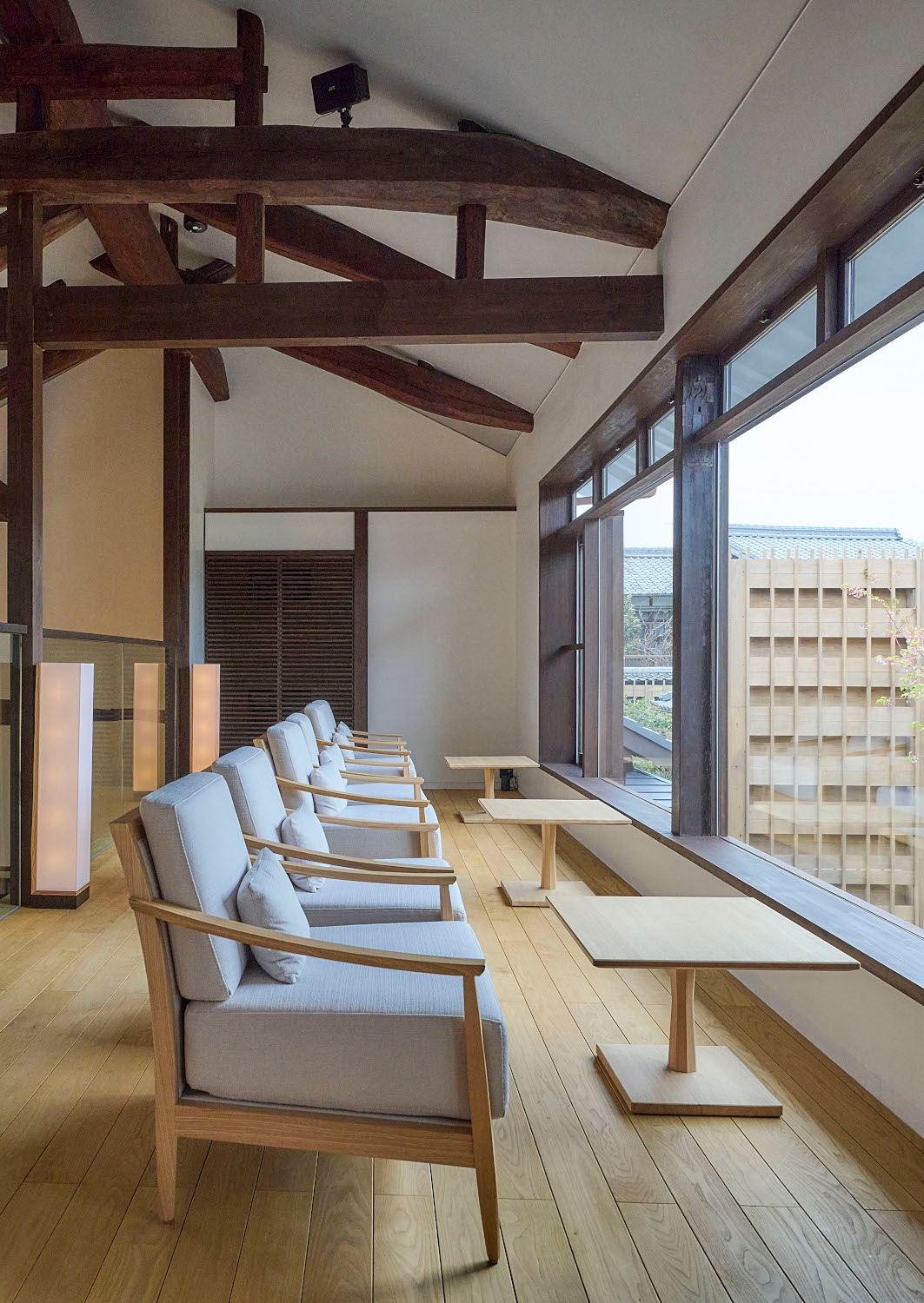 Interior of a lounge area with several white lounge chairs facing a few small wooden tables an a set of large picture windows looking out over buildings.