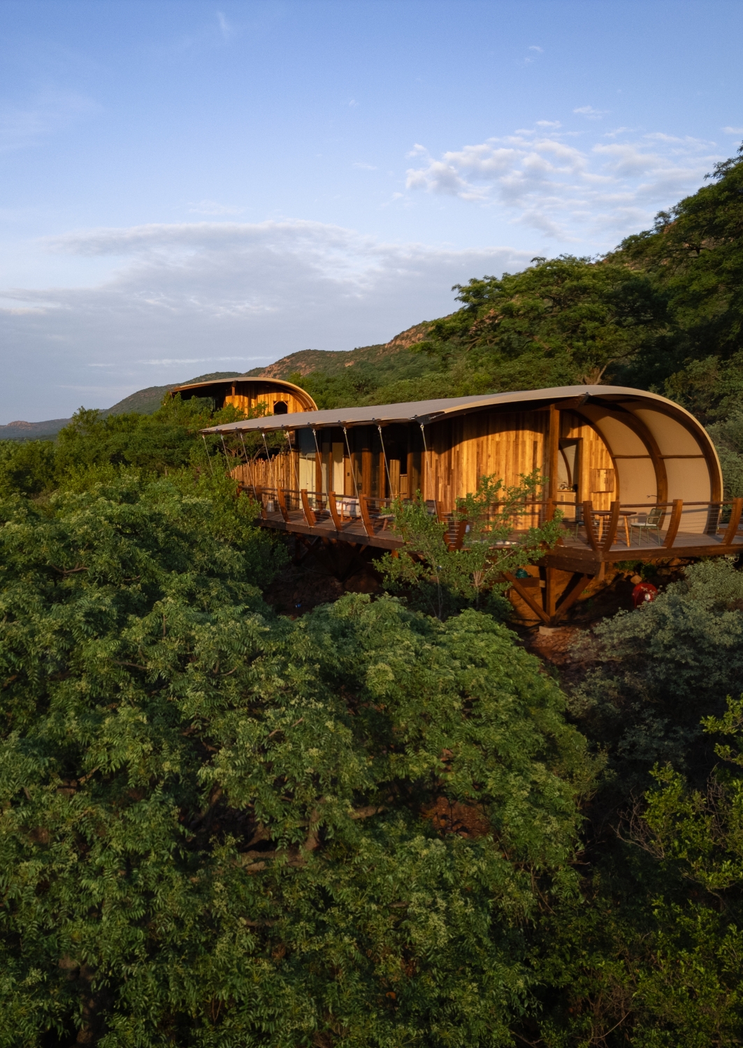 Exterior shot of two adjacent, cliffside safari suites with with canvas roof and wraparound wooden deck set among green trees with mountains in the distance.