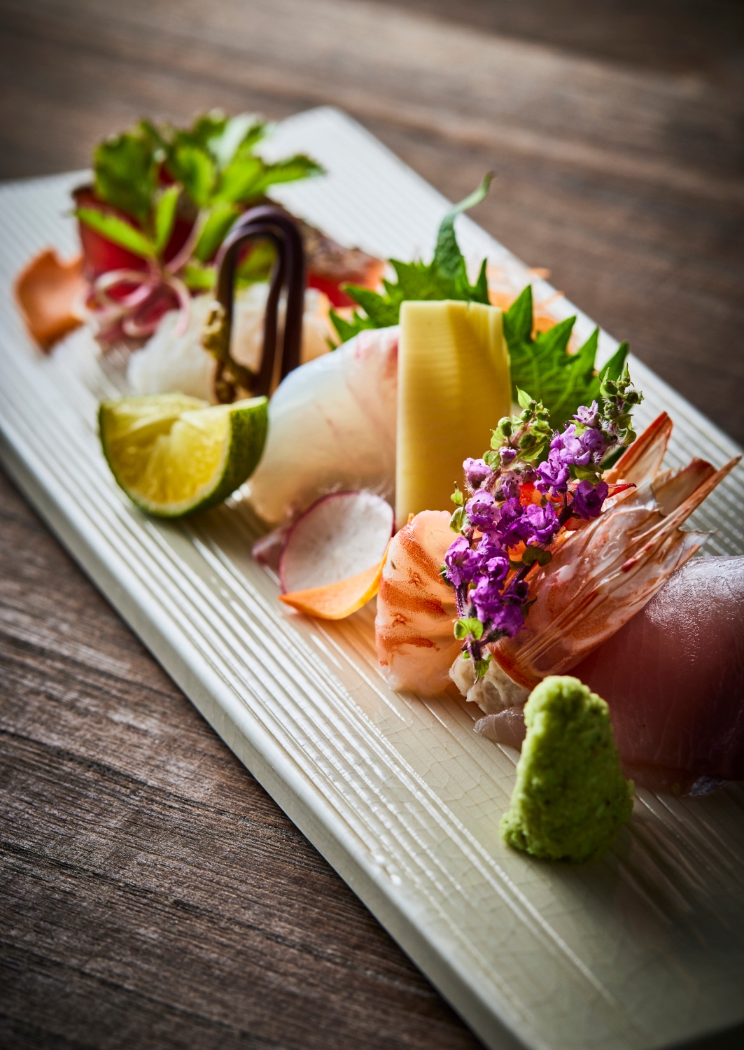 Wooden serving board with a selection of sushi presented with colourful flowers, leaves, limes and wasabi.