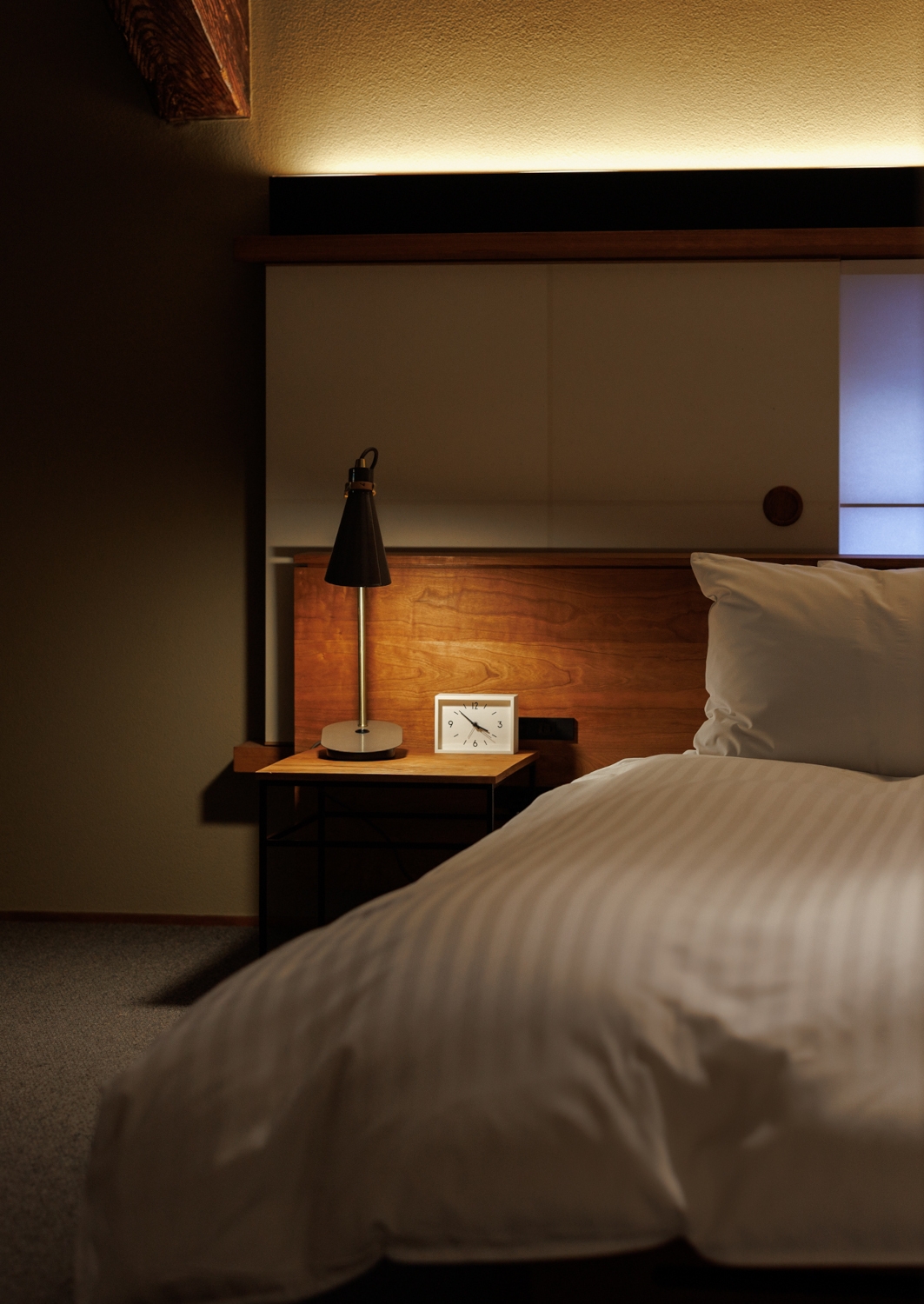 Interior of a ryokan berdroom with a western-style bed with white bedding and a small minimalist bedside table with a lamp on.