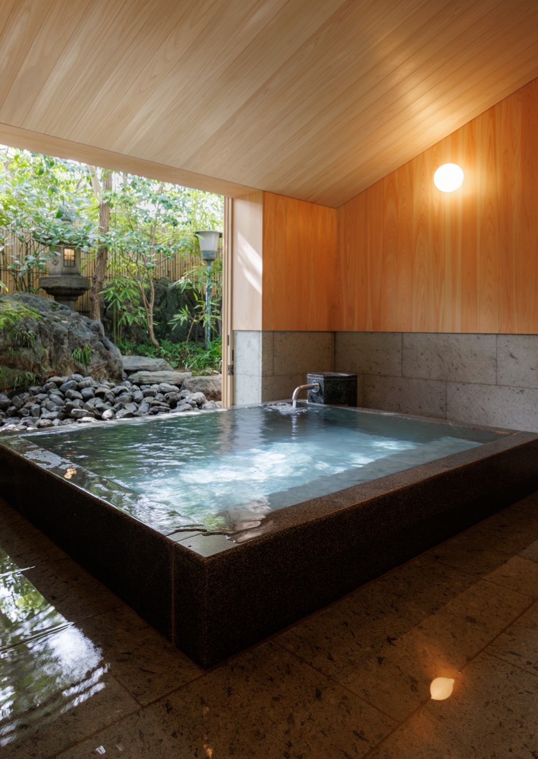 Traditional square Japanese stone onsen bath inside a wooden room with large windows looking out onto a garden