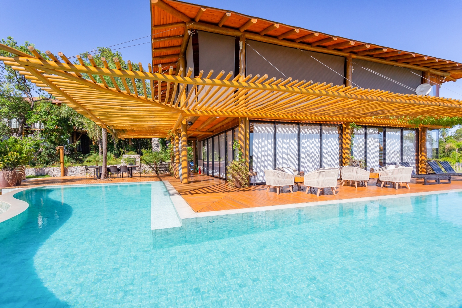 Exterior of the villa with an L-shaped wraparound pool in the foreground, wooden shading and the two-story villa and decking behind.