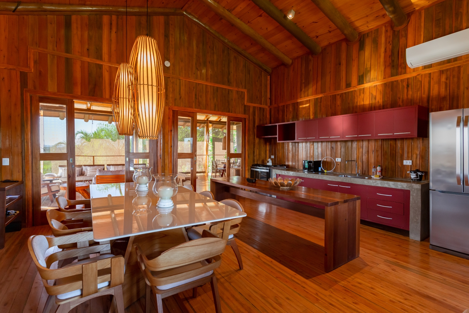Interior of the Bonito Villa kitchen - a full wooden interior with dining table in the foreground, large hanging light fixtures and a galley kitchen area.