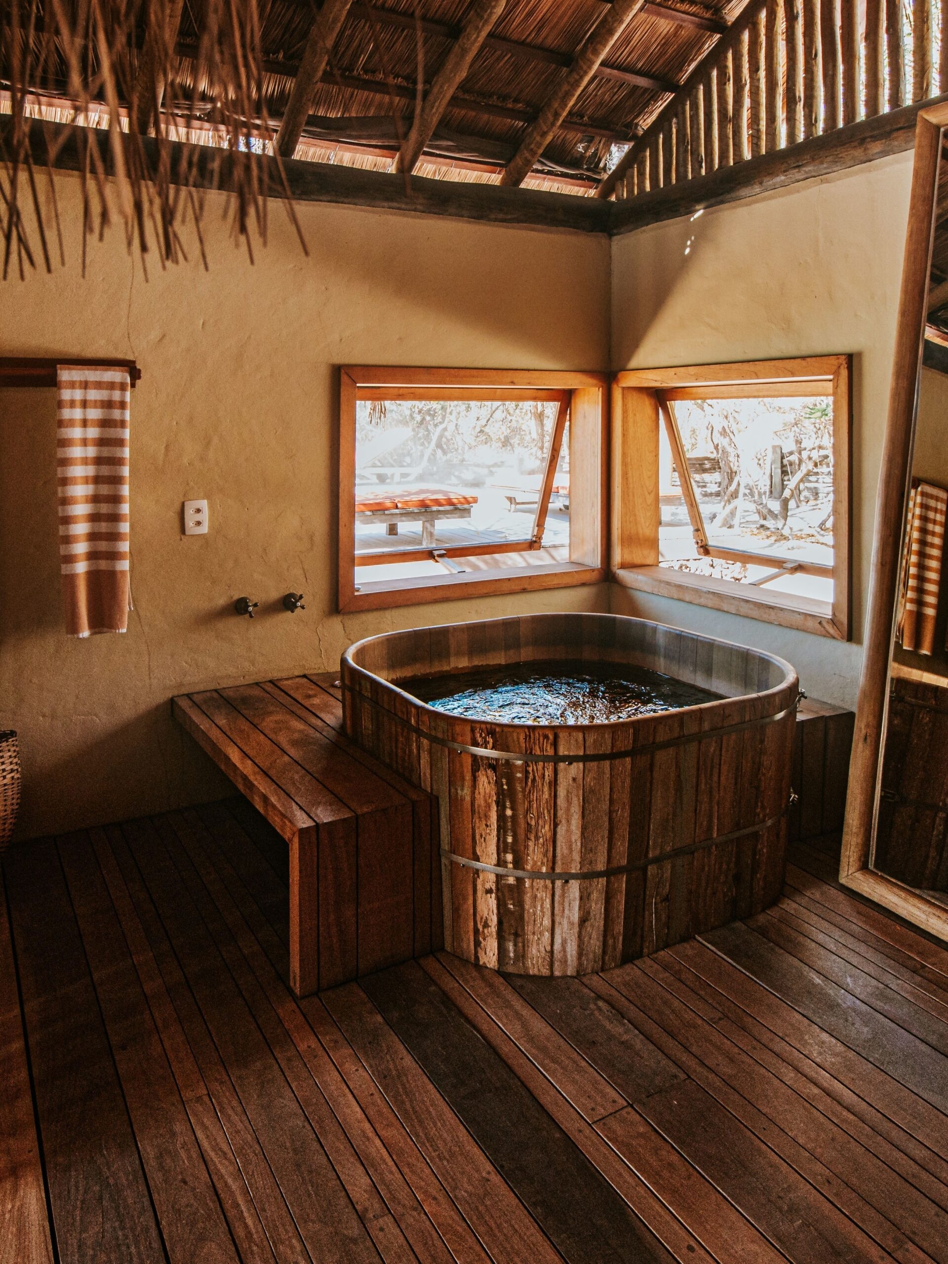A wooden hot tub with a bench and wooden floors in the same dark colour, in front of two windows and below a thatched roof.