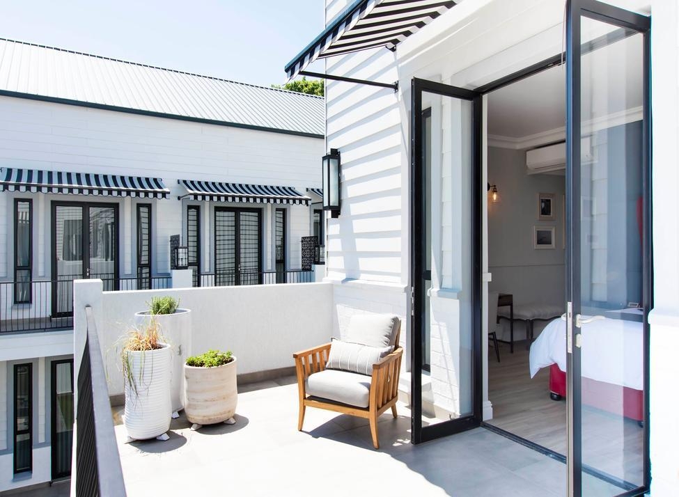 Glass doors opening out from a hotel suite to a large terrace with low stone walls, three potted plants and a chair with cushions. A bed is visible inside, and the terrace looks out to other hotel buildings, painted white with black and white awnings.