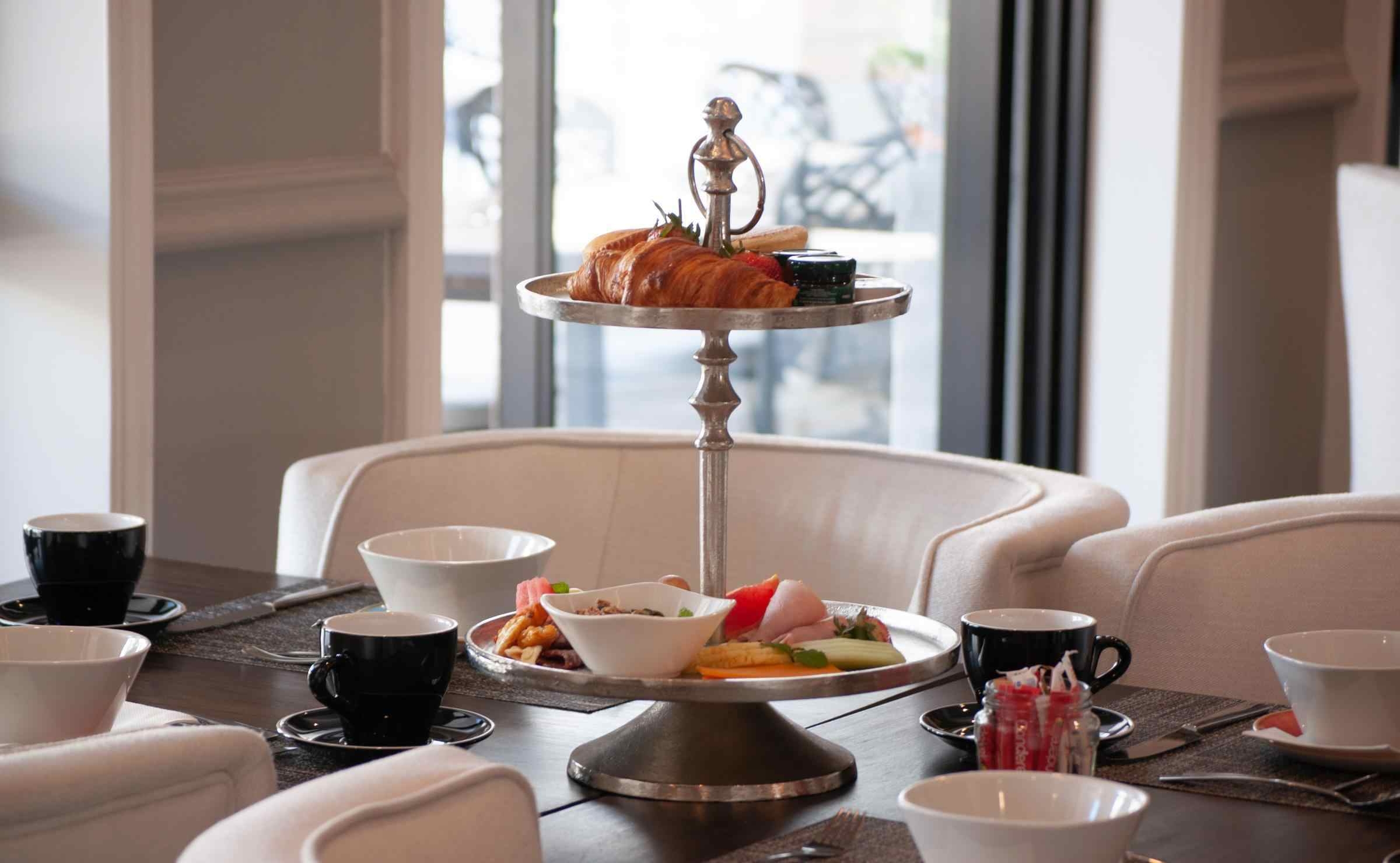 A close-up view of a table set for breakfast, with cream armchairs, cups and saucers and a cake stand with croissants, fruit and jams. Glass doors in the background lead to a terrace.