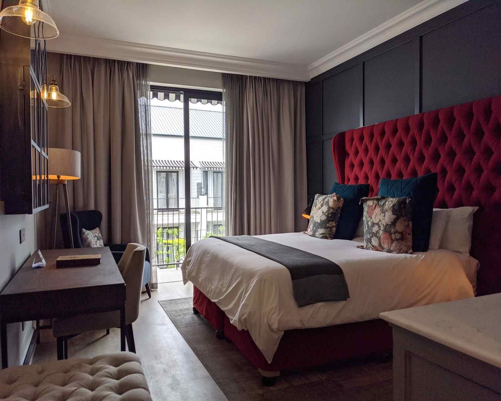 A dimly lit hotel bedroom with red headboard and partly drawn curtains showing views across a courtyard to a white building. There's a double bed, several lamps and a wooden desk with chair.