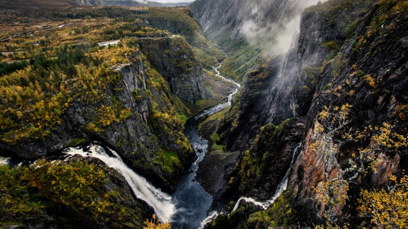 A dramatic deep canyon with a winding river and waterfall surrounded by steep, mossy rock walls and mist.