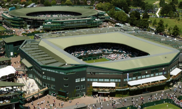 Aerial view of the green-roofed tennis stadiums at the All England Club in Wimbledon with surrounding spectators.