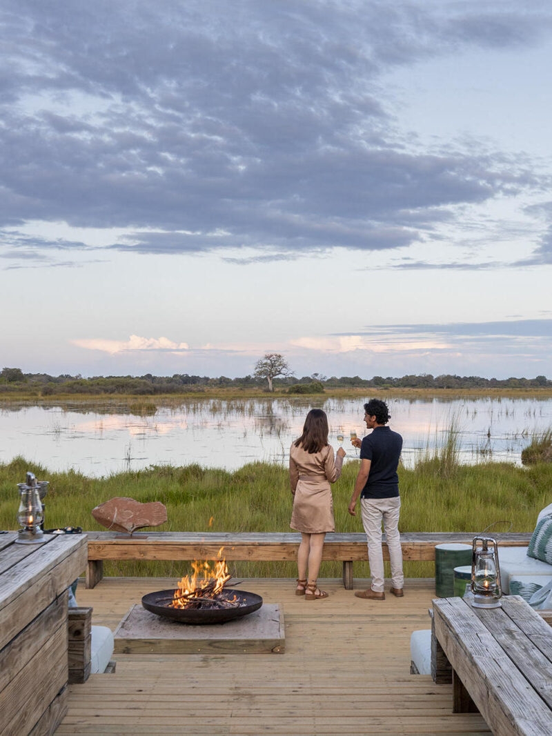 A man and a woman in a soft brown dress stand at the edge of a wooden verandah in front of a campfire, looking out over a still watering hole