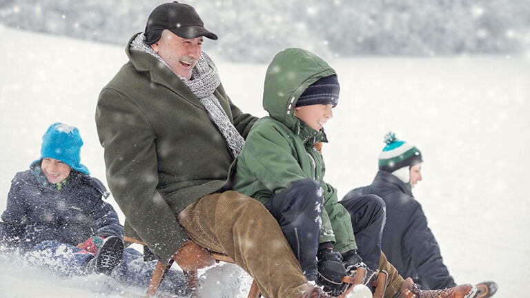An older man and children laugh while riding sleds down a snowy hill on their luxury Family holidays.