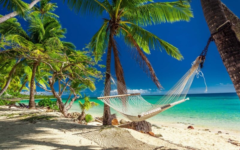 Rope hammock tied to palm trees on a white sand beach overlooking a tropical blue sea for luxury Fiji vacations.