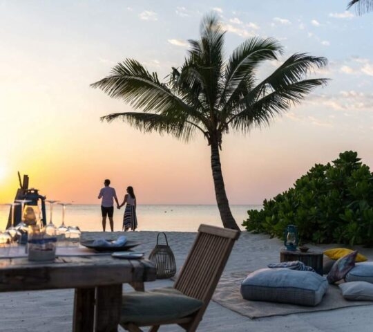 A table and beanbags are set up on the beach. At sunset, a couple can be seen holding hands.
