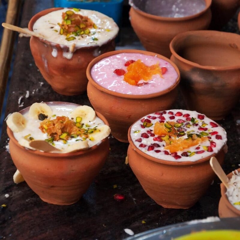 Four clay pots containing lassi topped with bananas, pistachios, and pomegranate seeds on a dark surface.