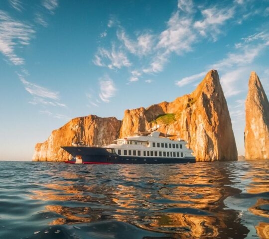 Luxury yacht sailing near large sunlit rocky cliffs protruding from the sea.