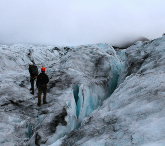Two hikers near a deep crevasse on a glacier.