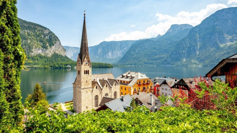 Church spire and colourful houses by a lake with mountain backdrop.