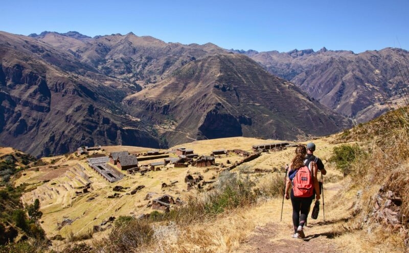 A hiker with a red backpack walking on a trail towards an ancient mountain village with terraces.