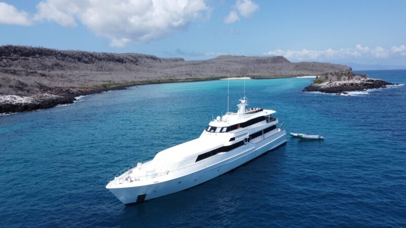 A luxury yacht anchored near a rocky coastline with turquoise waters, vegetation, and a small boat nearby in a sunny, scenic landscape.