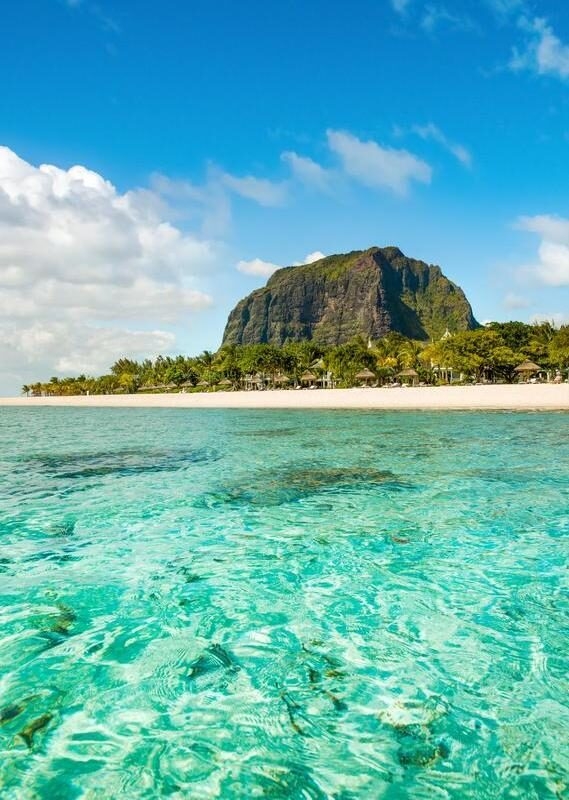 Eye-level view of a clear turquoise ocean with a sandy beach, tropical trees, and a large mountain in the background.