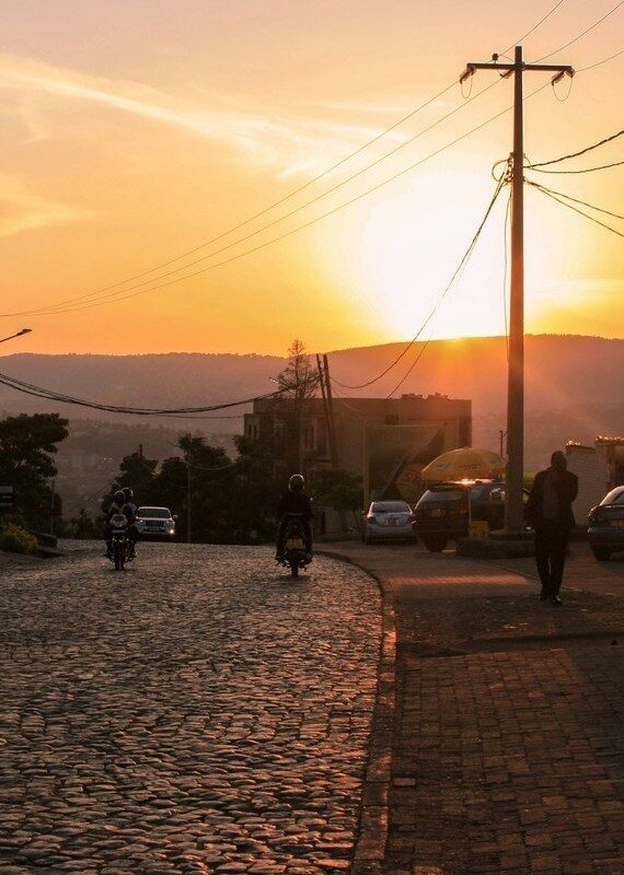Silhouetted commuters on a cobblestone street in Kigali during a golden sunset with a utility pole.