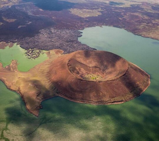 Aerial view of a large brown volcanic crater standing next to a green lake with rocky terrain.