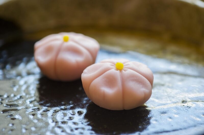 Two delicate pink flower-shaped sweets with small yellow centers placed on a textured dark ceramic plate.