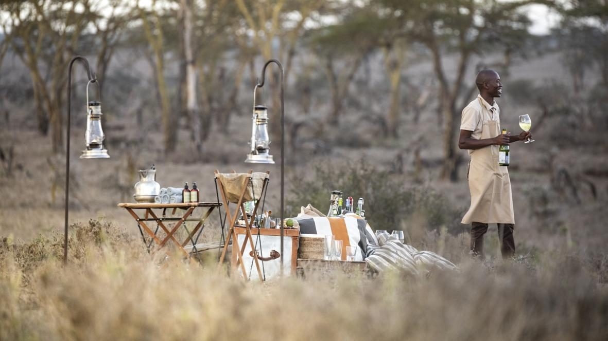 A staff member in an apron holding a wine glass beside a safari picnic setup with hanging lanterns in a field.