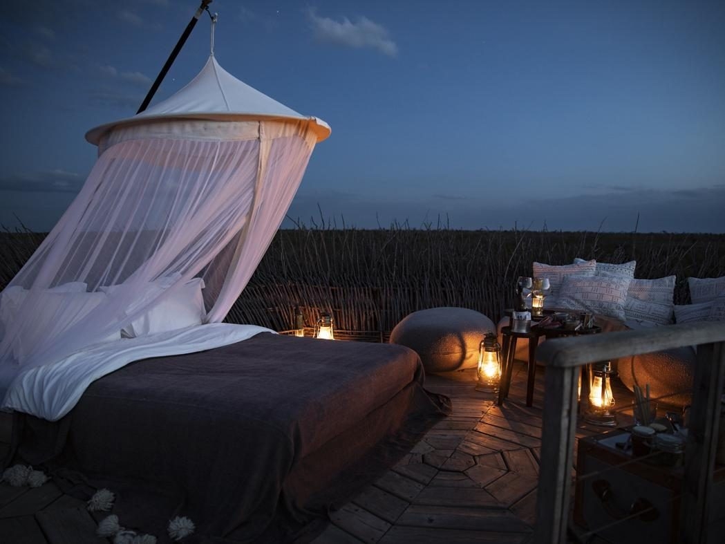 Open-air wooden deck featuring a bed with white canopy netting, surrounded by lit lanterns at dusk.