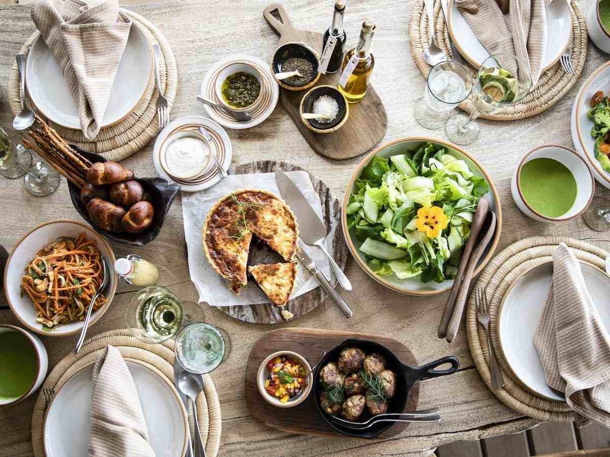 Top-down view of a wooden dining table spread with fresh green salad, quiche, bread rolls, meatballs, and wine glasses.