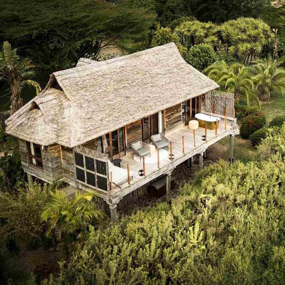 Aerial view of a wooden villa with a thatched roof, balcony seating, and an outdoor bathtub amidst trees.