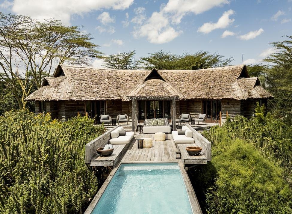 Thatched-roof villa with a wooden deck, outdoor lounge furniture, and a long swimming pool surrounded by trees.