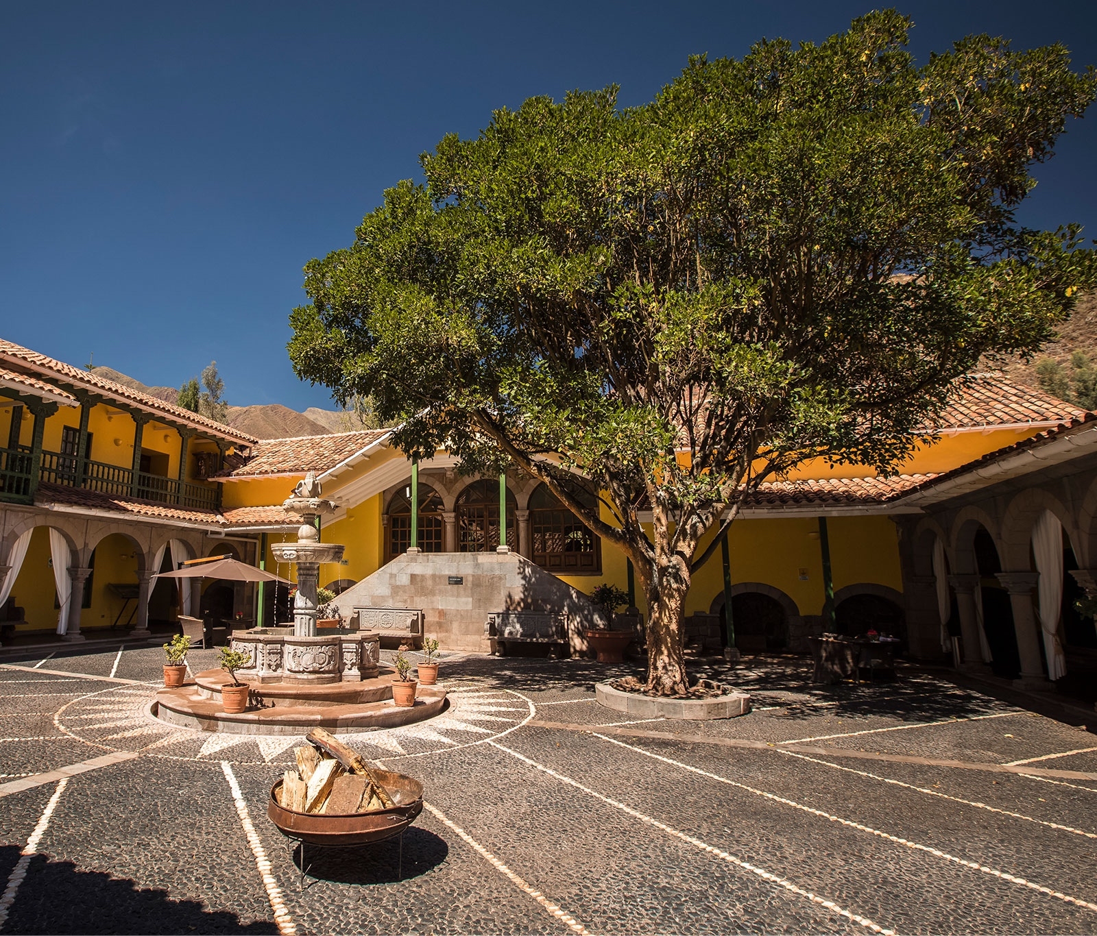Courtyard with a fountain, large tree, and cobblestone patterns, surrounded by yellow colonial buildings.