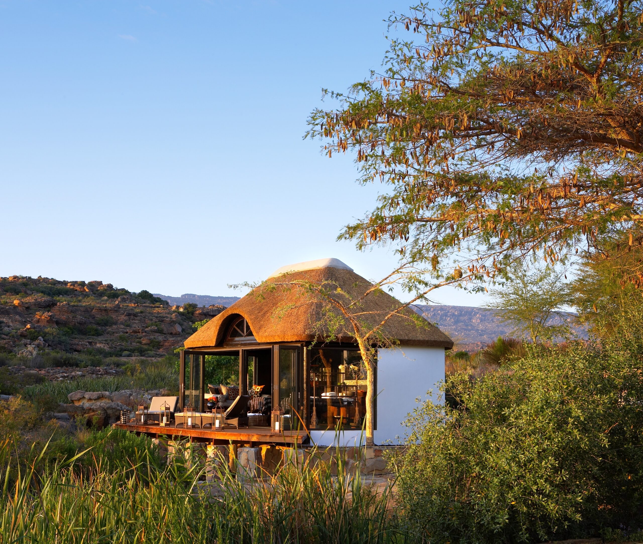 Thatched-roof safari lodge with glass walls nestled in a rugged, bushy landscape under a clear sky.