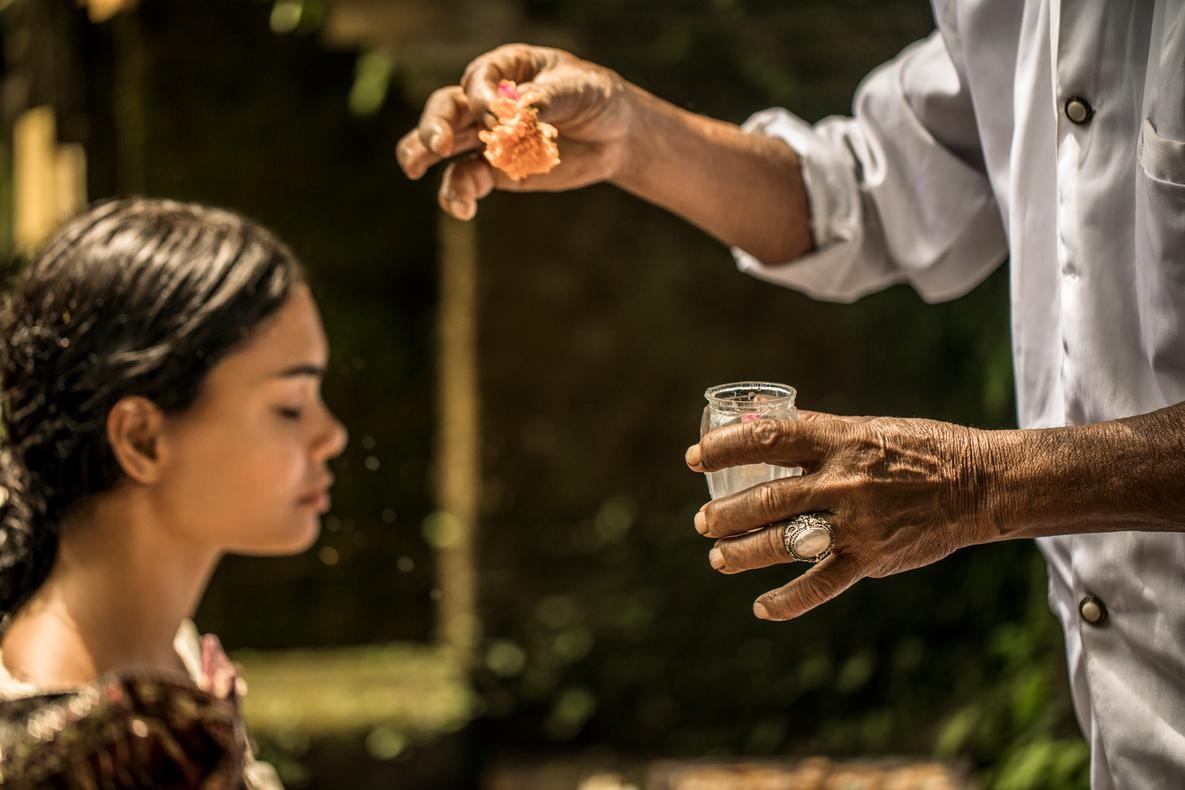 A close-up of a hand holding a flower and glass jar, sprinkling water onto a person with closed eyes.