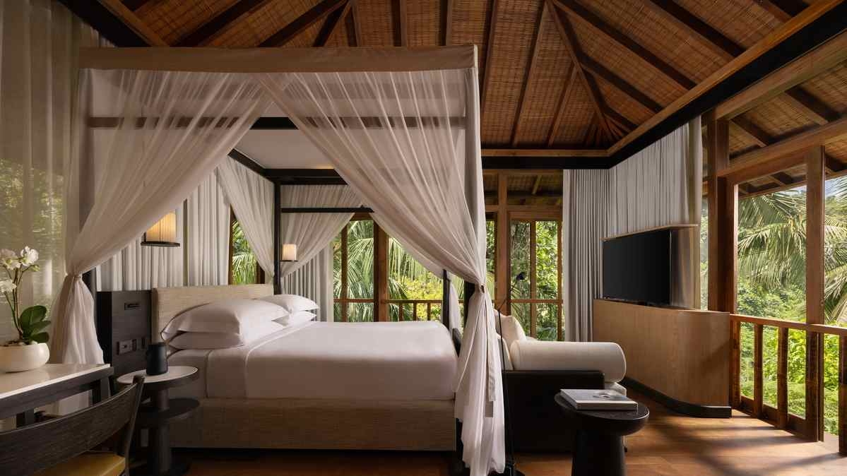 A bedroom with a four-poster canopy bed, wooden ceilings and large windows looking out to tropical greenery.