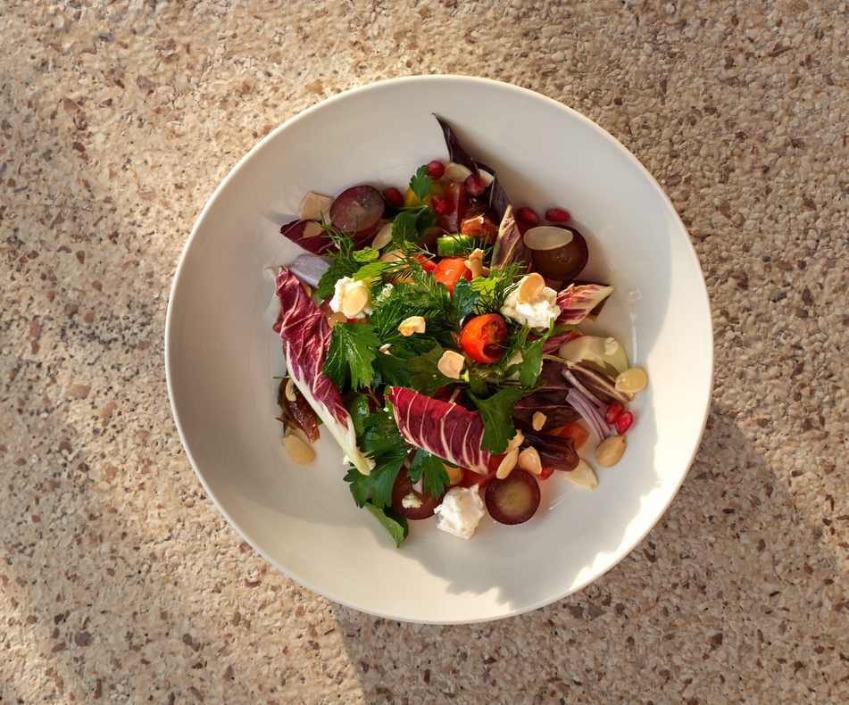 A white bowl filled with a fresh salad of green herbs, purple radicchio, grapes, cheese and almonds on a stone surface.