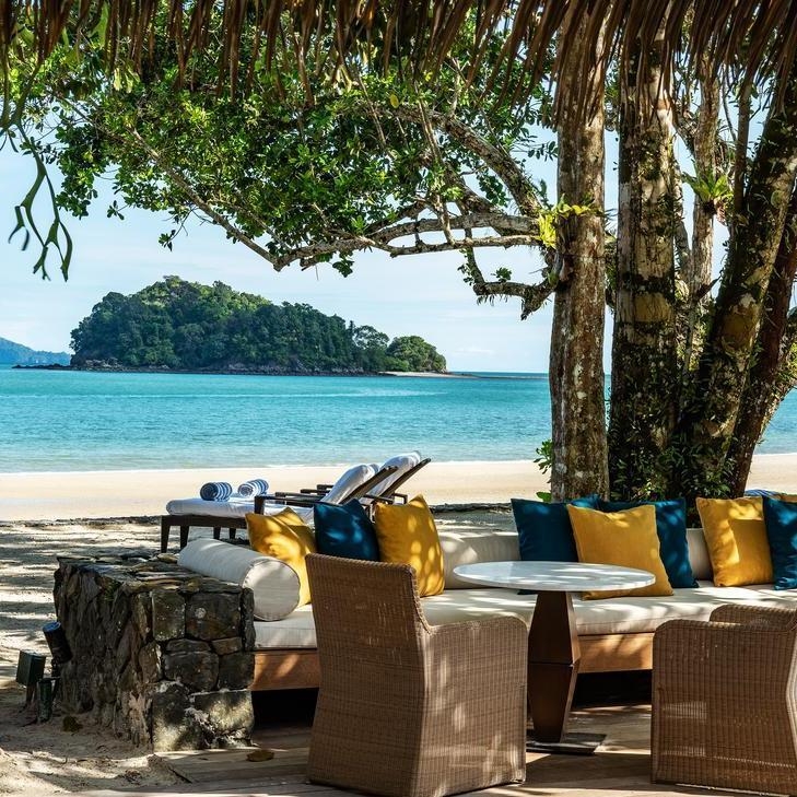 Shaded beach lounge with sofas and wicker chairs looking out at a tropical island in the sea.